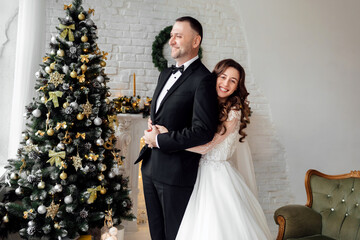 Young couple in love bride and groom posing in studio on background decorated with Christmas tree in their wedding day at Christmas. Enjoy a moment of happiness and love.