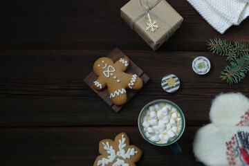 Christmas New Year card.Cup of hot drink with marshmallows, warm white mittens, New Year's gift, gingerbread, snowflakes, candles on a dark wooden background. Festive concept. Top view. Copy space.