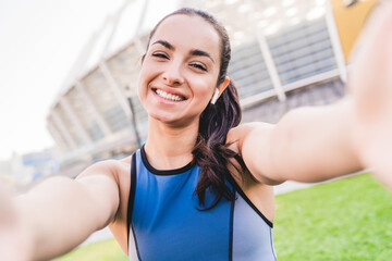 Fit caucasian female runner smiling while taking selfie wearing sporty top and earbuds with modern stadium in the background