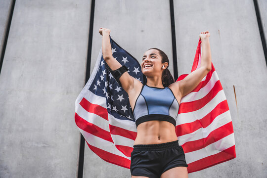 Low angle view photo of 20s happy female athlete holding American flag against grey wall