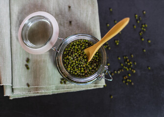 Top view of a glass container with mung beans. Transparent pot open on a tablecloth full of vegetables with a wooden spoon.