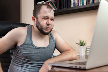 Leisure using laptop. A young bearded guy in home clothes gray T-shirt, uses laptop for watching...