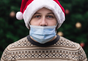 Wearing a mask incorrectly. Close up Portrait of man wearing a santa claus hat, xmas sweater and medical mask with emotion. Against the background of a Christmas tree. Coronavirus pandemic