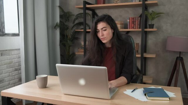 Young pensive woman employee in the office at the computer. Brunette girl fills out a questionnaire or resume, takes a test on a laptop. Work during self-isolation and quarantine COVID-19 4k