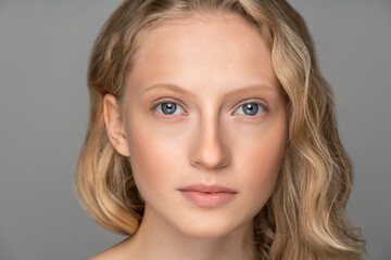 Close up of young woman face with blue eyes, curly natural blonde hair and eyebrows, has no makeup,...