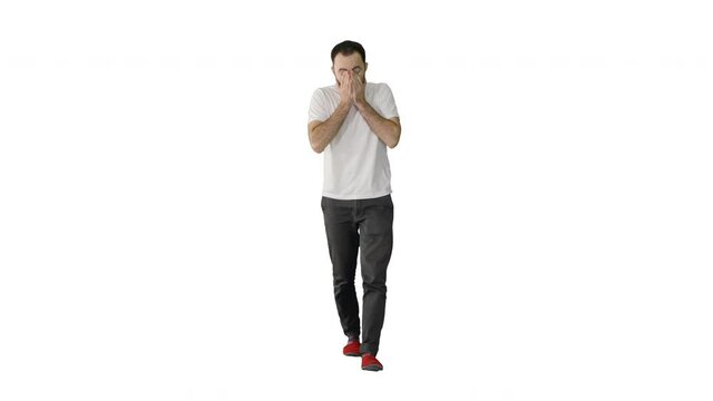 Sleepy male in white t-shirt yawning and rubbing eyes while walking on white background.