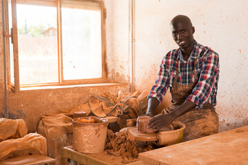 Smiling African American guy enjoying work with clay on potter wheel in ceramics workshop