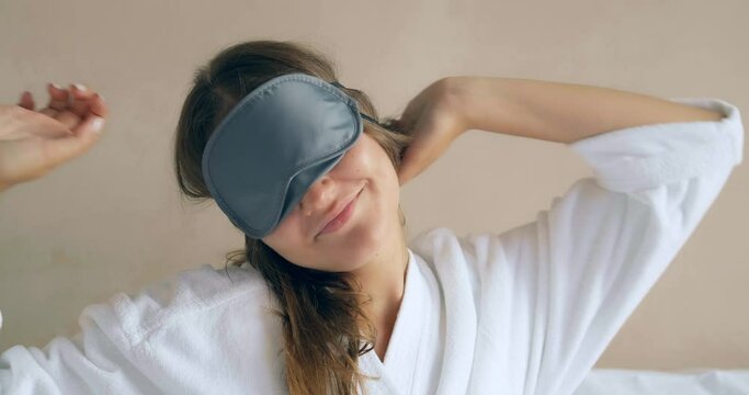 smiling woman in white bathrobe takes off black sleep mask sitting against wall in hotel room in morning closeup