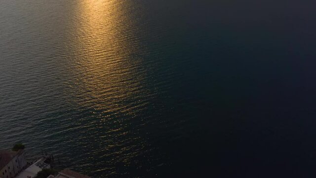 Slow, aerial, tilt shot revealing Lake Garda at sunset with warm sunset light braking through the clouds and spilling over the mountains