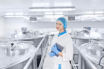 Young female worker operator in uniform holding computer tablet near food factory production line