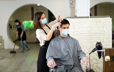 Female hairdresser making hair styling for young man in protective mask and gloves, working day during pandemic situation
