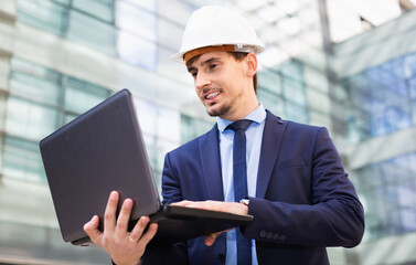 Positive smiling professional man in jacket and helmet working at electonic device outdoor