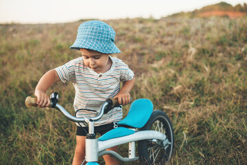 Obraz premium Caucasian small boy walking with a bike on a field wearing a blue hat in a summer day