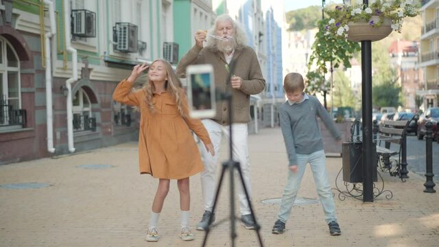 Joyful boy and girl dancing as senior man blowing soap bubbles. Portrait of cheerful multi generational family recording funny video for social media outdoors.