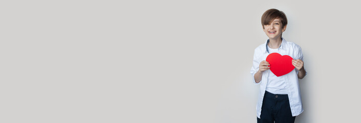 Young boy is holding a heart from paper and smile on a gray studio wall with free space in casual clothes