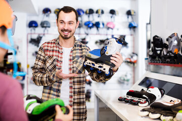 smiling spanish male shop assistant helping boy to choose roller-skates in sports store.