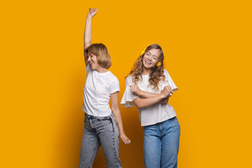 Blonde woman with her friend is listening to music on a yellow wall using headphones and dancing