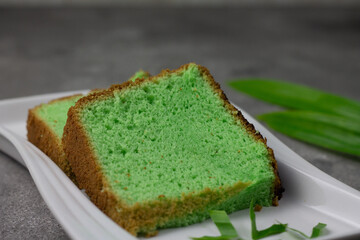 Bolu Pandan or Chiffon Pandan Cake served on ceramic white plate, with soft and moist textures. Garnished with pandan leaves on grey wooden textured background. Close up view, copy space. 