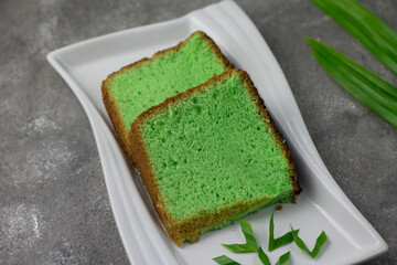 Bolu Pandan or Chiffon Pandan Cake served on ceramic white plate, with soft and moist textures. Garnished with pandan leaves on grey wooden textured background. Close up view, copy space. 