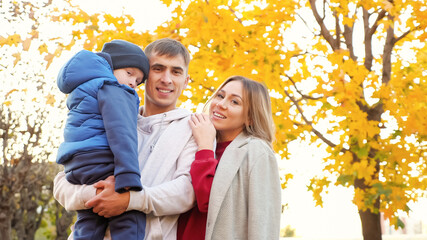 Lovely family of mother father and funny little boy hug posing for camera in picturesque autumn city park on nice warm day low angle shot