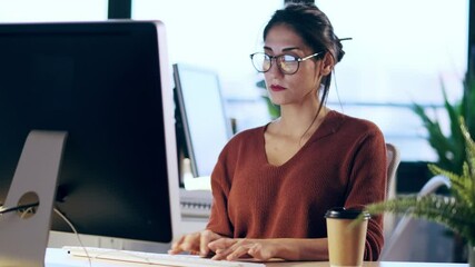 Shot of beautiful young business woman working with computer while talking with earphone sitting in the office. - Powered by Adobe