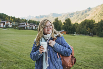 Young blonde woman in jeans coat with paper cup of morning coffee at the chalet cottage village on...