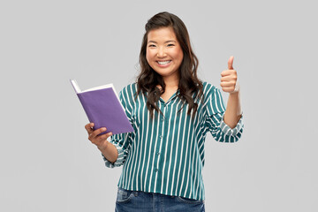 people, ethnicity and portrait concept - happy asian young woman with book showing thumbs up over grey background