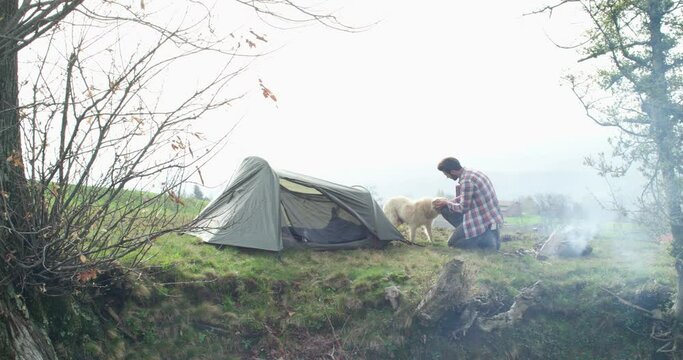 Young hipster man with man's best friend. Man with his dog camping near bonfire, in front of tent. Grey sky in background 