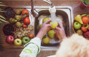 Hands of an elderly man washing a yellow apple under running water. Table full of different fruit for a healthy lifestyle