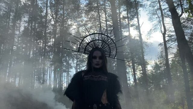 Woman in image of witch walks in penetrated by the sun rays foggy forest.