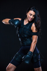 A young athletic woman in an EMS suit with Boxing gloves on an isolated black background. EMS training. Electro muscular stimulation machine.