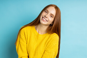 Fototapeta premium Attractive excited happy woman with red long hair, freckles, no make up smiling cheerfully showing her perfect white teeth, wearing yellow sweter posing on blue background.
