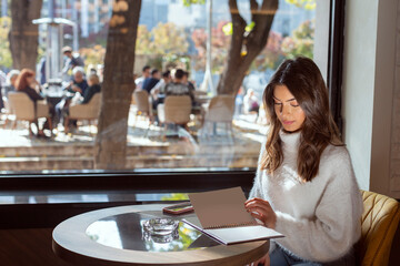 Beautiful young woman, alone, looking at the menu, deciding what to order in the restaurant