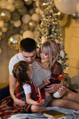 Mom, Dad and Daughter reading Magic Book At Home Near Chritmas Tree. Celebration Atmosphere. Magic Lights In Night Xmas Tree Interior. Lollipop. 