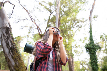 Caucasian young lady shooting landshate and trekking in woods. Female traveler exploring nature, holding camera, taking photo and carrying big backpack. Tourism, adventure and summer vacation concept