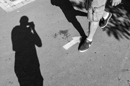 a shadow of a street photographer sealing the legs of a man walking against a painted arrow. street black and white photography.