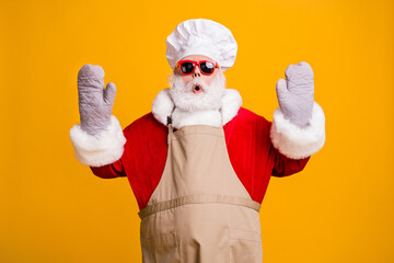 Photo of confused santa grandpa chef headwear bake cookery raise hands gloves open mouth tried help did wrong move wear red costume coat sun specs cap apron isolated yellow color background