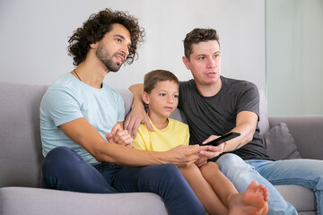 Excited homosexual dads and son watching TV show at home, sitting on couch in living room, hugging, using remote control, looking away. Family and home entertainment concept