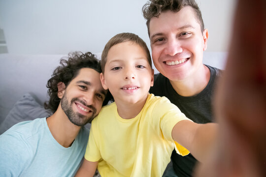 Selfie Of Cheerful Homosexual Dads And Kid Sitting On Couch At Home And Smiling At Frontal Camera. Front View. Family And Communication Concept