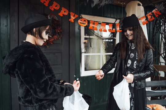 Scary girls, sisters, friends celebrating halloween. Playing treak or treat game on porch with garland. Bags with sweets in hands.Terrifying face skull makeup.Witch stylish costumes. Children's party