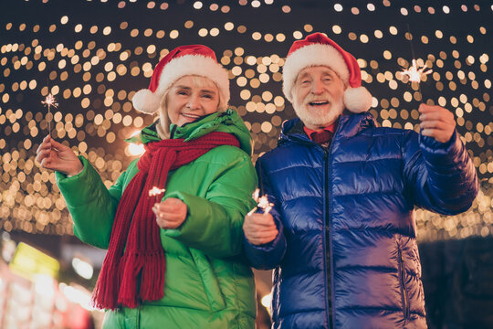 Photo of two people pensioner couple man woman meeting hold bengal fire cheerful smile go main square evergreen tree wear coat red scarf headwear evening street illumination fair outside