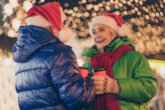 Photo of two people retired man woman meeting hold cup drink mulled-wine laugh x-mas joke comfort relax wear coat headwear mittens red scarf evening street illumination fair outdoors