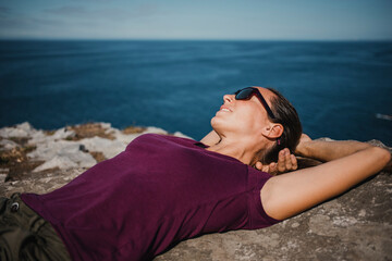 Naklejka na ściany i meble Lying woman on a rock, at the coast line with sunglasses and purple t shirt