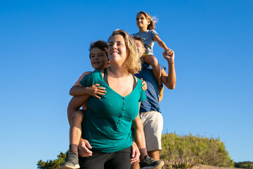Obraz premium Happy family enjoying hiking in countryside. Two kids riding on parents back and neck. Low angle. Nature and recreation concept