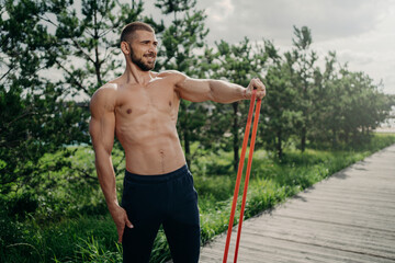 Strength and motivation concept. Muscular man with naked torso, has exercises with resistance band, performs training for biceps, looks into distance, poses outdoor. Sportsman uses expander.