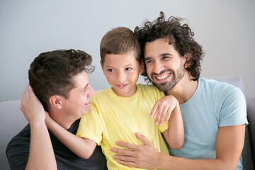 Cheerful gay fathers and son sitting on couch together and hugging each other. Front view. Happy family and parenthood concept