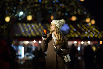 a blonde girl, in the evening, against the background of the lights of the fair with a drink in an orange glass, a white hat and a beige coat walks on the street.