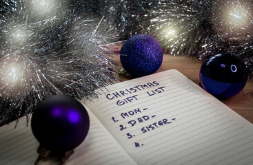 Blue Christmas baubles, a notepad and dim lights. An English text handwritten in a paper notebook "Christmas gift list - mom, dad, sister".