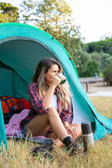 Caucasian young woman drinking tea, sitting in tent and looking away. Thoughtful female traveler...
