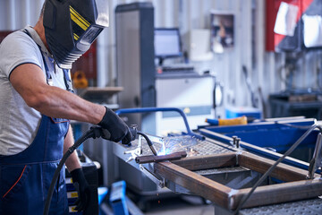 Mechanic in protective helmet welding metal car parts in garage